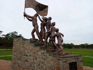 A wide shot of the public Independence bronze monument located at Kololo Ceremonial Grounds in Kampala. The monument stands on a tiered stone base set against a vast green field and a grey sky.