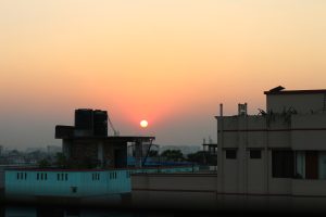 A serene sunset view over a cityscape, with the sun partially visible just above the horizon. In the foreground, roofs of buildings are silhouetted against the sky, which transitions from orange to a soft blue gradient.
