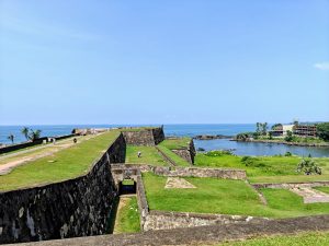 Stone ramparts of Galle Fort, Sri Lanka, with grassy terraces, walking paths, and arched passages overlooking the Indian Ocean under a clear blue sky.
