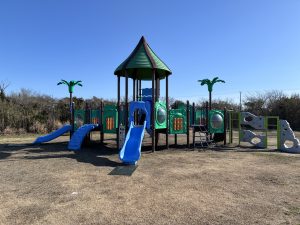 
A playground structure featuring a green and brown color scheme, with a peaked roof and palm tree-like decorations. It includes blue slides, climbing areas, and various interactive panels.