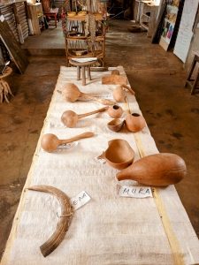 Traditional wooden and gourd artifacts from Bugisu displayed on a white woven mat, including gourds, containers, and a small three-legged stool.