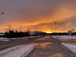 A snowy street at sunset, with warm orange light glowing across the sky and reflecting on the icy road