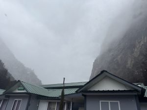 A gray hotel building with a green metal roof is partially visible in the foreground. Behind it, a steep, rocky mountain is partially obscured by clouds and fog, creating a misty atmosphere.