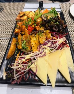 A long black serving platter filled with a variety of vegetarian appetizers, including grilled paneer cubes, seasoned potato wedges, sautéed vegetables, and a dark, glazed black mushroom. The platter is garnished with fresh shredded purple cabbage and sprouts, with three triangular slices of cheese arranged at the front.