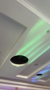 Modern white ceiling with a circular air vent featuring a black grille, illuminated by soft green and blue light streaks with recessed lighting nearby.
