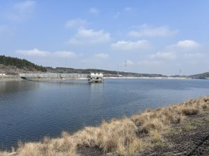 Calm water with grassy shoreline, a small stilted building, and a dam under a cloudy sky at Ichizu Lake, Nagara.
