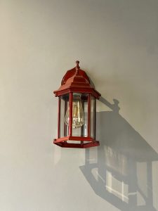 A vintage-style red metal lantern with clear glass panels mounted on a light gray wall, casting a long shadow to the right.
