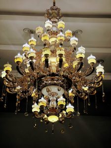 A grand, multi-tiered ornate chandelier hangs from a ceiling, featuring warm, glowing candle-shaped bulbs and elegant alabaster-style shades. The gold-finished frame is decorated with intricate scrollwork and dangling crystal droplets that catch the light.