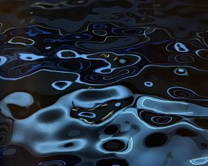 Close-up of a highly polished, reflective metal surface designed with a sculptural rippling pattern that mimics the look of dark water. Glossy distorted reflections of light in dark blue, cyan, and black create organic circular and wavy shapes across the textured surface