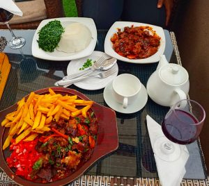 Glass table with stir-fried goat meat, fries, kachumbari, posho, greens, and drinks including tea and red wine.