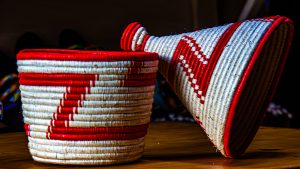 A handcrafted basket with a conical lid, woven in red and white patterns, placed on a wooden surface.