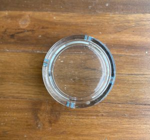 A top-down, centered photograph of a clear circular glass ashtray with four notches, resting on a rustic brown wooden tabletop with a visible natural grain.
