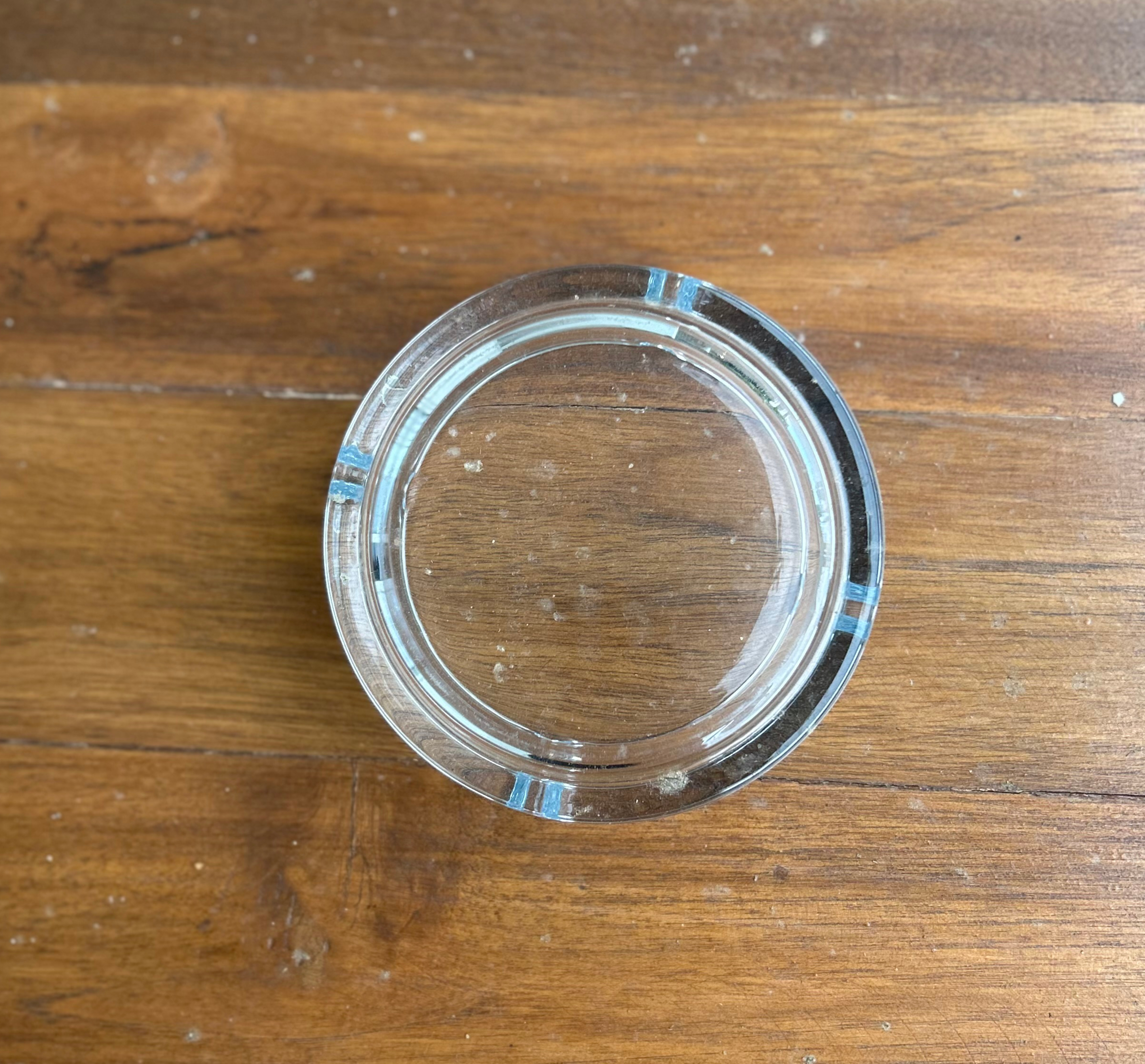A top-down, centered photograph of a clear circular glass ashtray with four notches, resting on a rustic brown wooden tabletop with a visible natural grain.