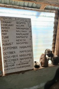 A vertical white board displaying the months of the year translated into local Eastern Uganda dialects. The board sits on a concrete windowsill next to small clay sculptures and a window covered in translucent plastic.