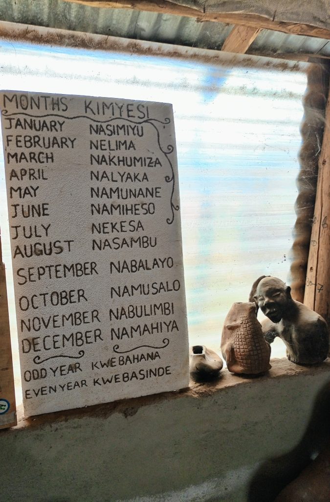A vertical white board displaying the months of the year translated into local Eastern Uganda dialects. The board sits on a concrete windowsill next to small clay sculptures and a window covered in translucent plastic.