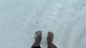 A person stands in shallow, clear sea water on a white sand beach, with feet visible through the water.