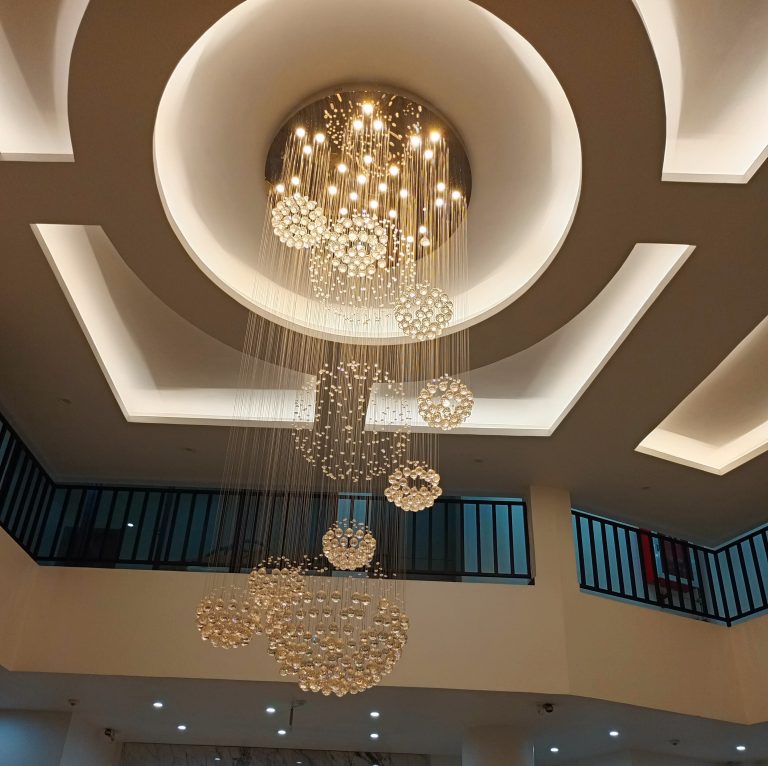 A low-angle view of a modern crystal chandelier with hanging spheres, set against a geometric recessed ceiling, with a balcony railing in the background.