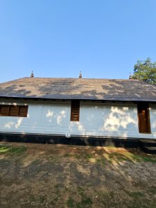 A traditional Kerala style temple building with a sloping metal tiled roof and white walls under a clear blue sky, featuring wooden windows and a door. Captured at Niramkaitha Kotta Temple, Vallikunnu North, Malappuram.