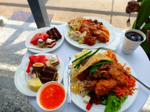 A high-angle view of a buffet on a white table at the Kigali Convention Center, with plates of rice, curry, grilled chicken, chapatti, vegetables, and desserts like cake and fruit.