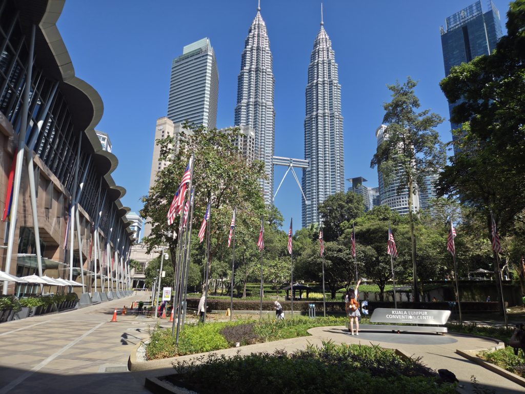 The image captures a sunny day in an urban park in Kuala Lumpur, featuring the iconic Petronas Twin Towers in the background.
