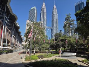 The image captures a sunny day in an urban park in Kuala Lumpur, featuring the iconic Petronas Twin Towers in the background.