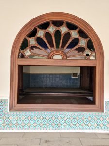 An elegant arched wooden window with decorative glass patterns, opening into a calm interior space. The tiled border adds charm in Kozhikode, Kerala. 