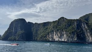 A long, towering limestone cliff face heavily covered in green trees, with various speedboats and longtail boats cruising in the vibrant blue sea.