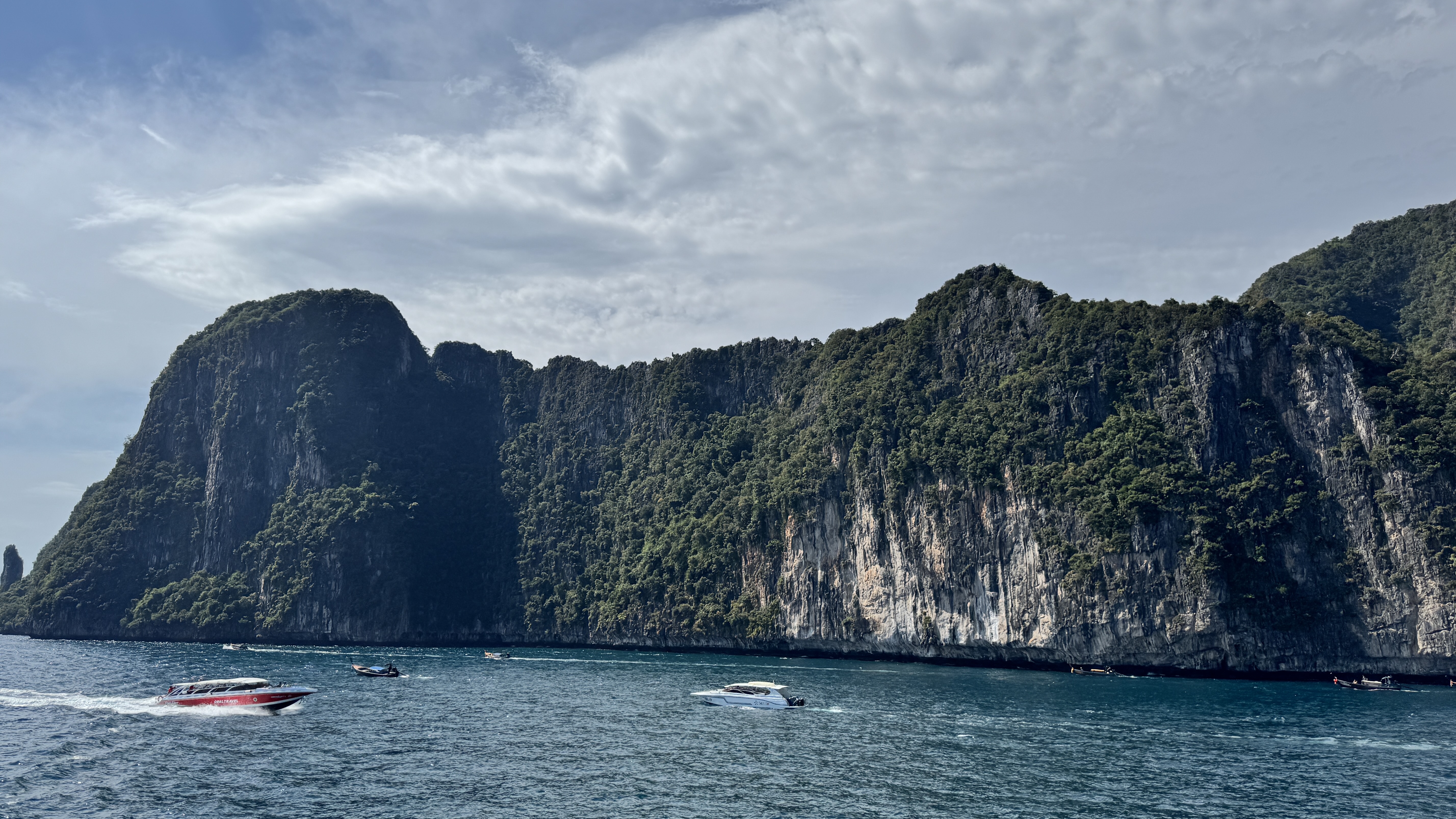A long, towering limestone cliff face heavily covered in green trees, with various speedboats and longtail boats cruising in the vibrant blue sea.