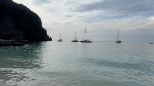 Several white sailing yachts and boats floating on calm tropical ocean water under a cloudy sky with a dark mountain cliff on the left side.
