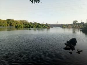 Wide landscape view of Nalubaale Dam and the Source of the Nile Bridge in Jinja, with calm river, rocks, and lush green banks under a clear sky.
