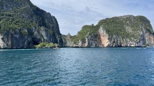 A wide landscape view of a massive limestone island with steep, jagged cliffs and dense green vegetation rising from the calm blue sea.