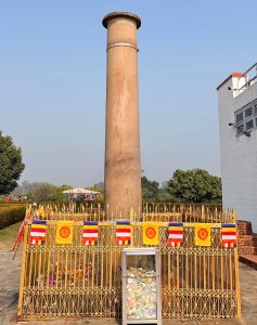 A tall, cylindrical pillar made of reddish-brown material stands prominently against a clear blue sky. The pillar is surrounded by a decorative golden fence adorned with colorful flags featuring various patterns, including circles and stripes.
