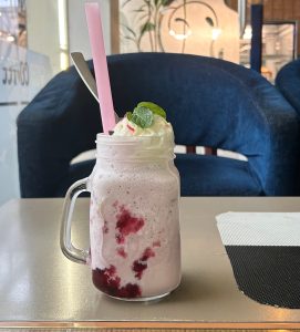 A transparent glass jar filled with a smooth, creamy pink drink. It is topped with soft whipped cream and a fresh green mint leaf, giving it a cool and refreshing look.
