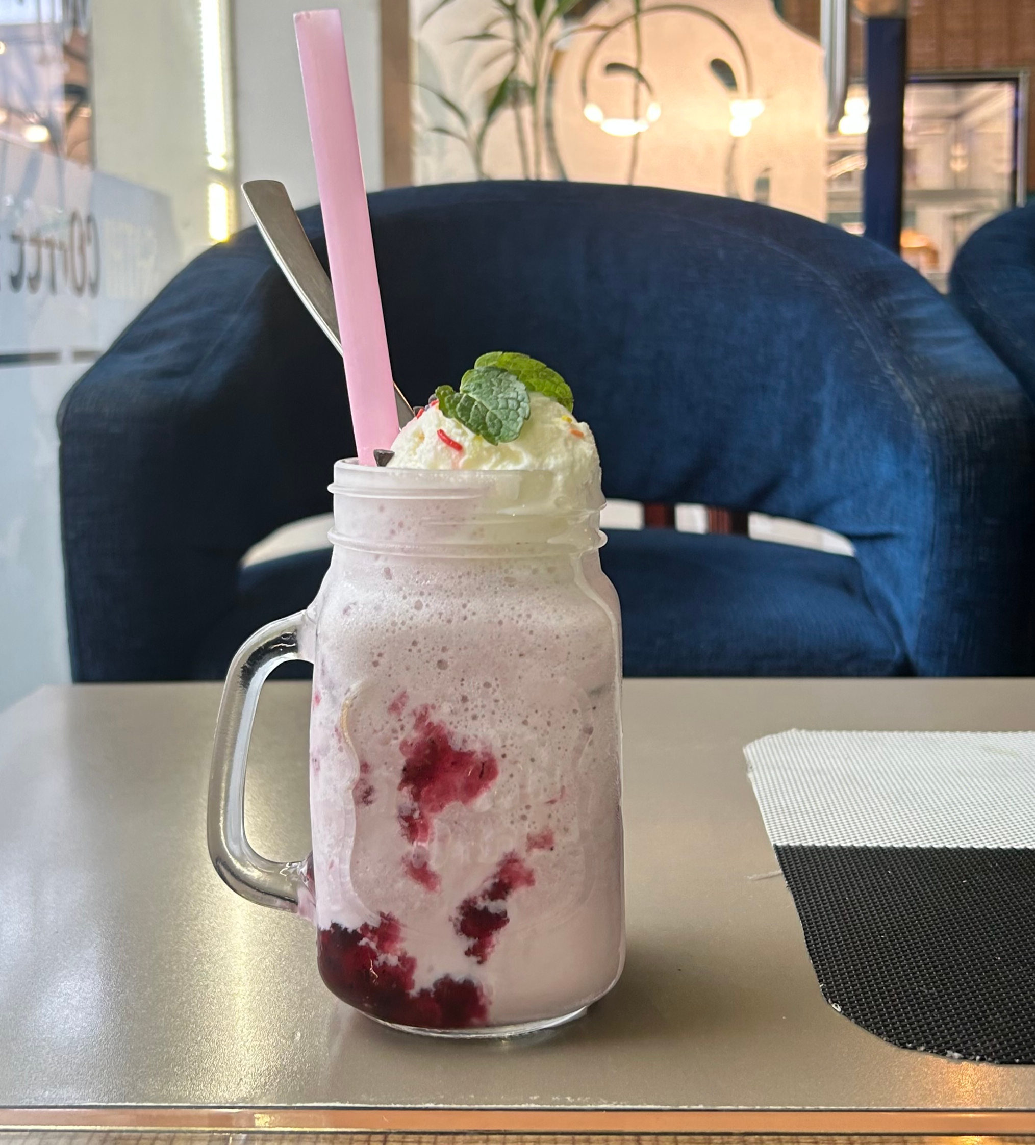 A transparent glass jar filled with a smooth, creamy pink drink. It is topped with soft whipped cream and a fresh green mint leaf, giving it a cool and refreshing look.
