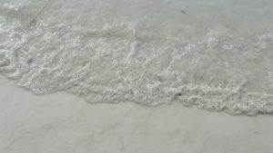 A close up shot of clear ocean water waves with white foam washing over a light colored sandy beach.
