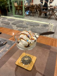Ice Cream Sundae, Vanilla ice cream scoop in a crystal glass bowl topped with chocolate and caramel drizzle and colorful star-shaped sprinkles. The dessert is served on a wooden coaster at a restaurant with a warm, blurred interior background.