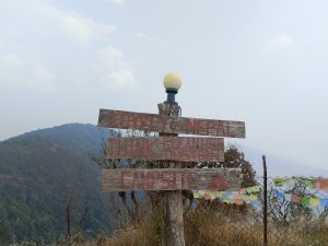 A rustic wooden signpost on a hillside reads “Lovely Nepal,” “Think Clean Use Dustbin,” and “Campsite,” with mountains and a cloudy sky in the background. Colorful prayer flags hang on a nearby fence.