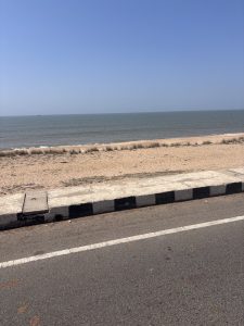 A tranquil scene with a sandy beach dotted with sparse grass, a calm blue ocean stretching into the distance under a clear sky, a narrow road with a white center line running alongside the shore, and a small boat visible far out on the water.
