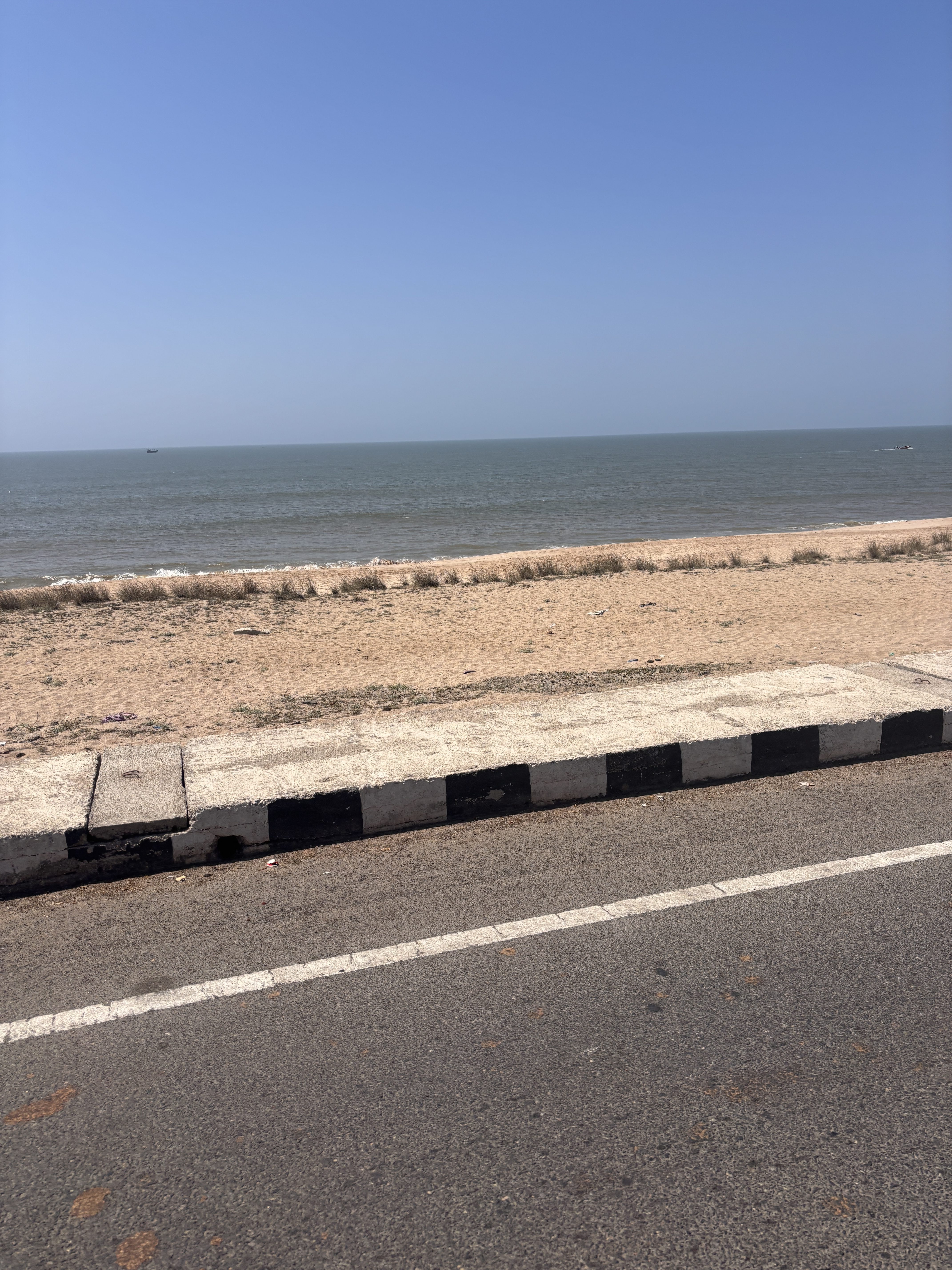 A tranquil scene with a sandy beach dotted with sparse grass, a calm blue ocean stretching into the distance under a clear sky, a narrow road with a white center line running alongside the shore, and a small boat visible far out on the water.