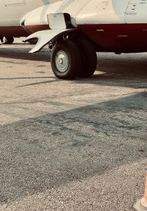 A close-up shot of the dual-wheel main landing gear of a white and red commercial aircraft on a sunlit asphalt tarmac. The image captures technical details of the tires, hydraulic struts, and open gear bay doors.