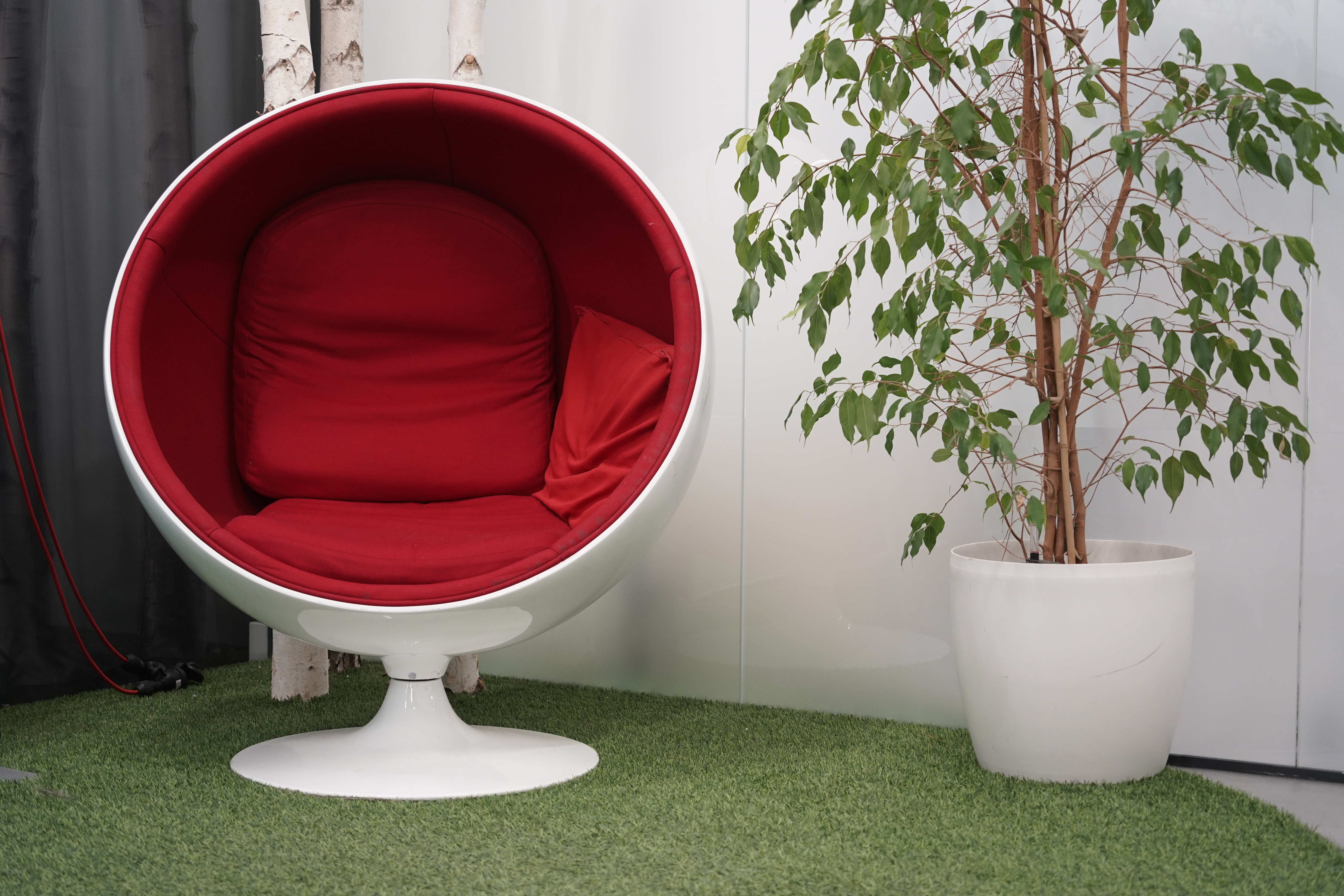 A modern, circular chair with a deep red cushion is positioned on a green, grassy carpet. The chair has a smooth white exterior and a wide base, giving it a unique, pod-like appearance. Adjacent to the chair is a potted plant with slender brown branches and numerous small, green leaves