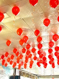 Dozens of vibrant red helium balloons with curling gold ribbons floating against a decorative white textured ceiling. The perspective is looking upward, creating a festive and celebratory atmosphere.
