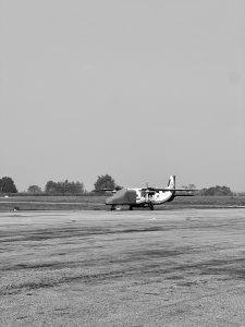 A black and white photograph of a small twin-propeller aircraft parked on an airport tarmac. The nose of the plane is covered with a protective tarp, set against a flat horizon with distant trees under a clear sky.