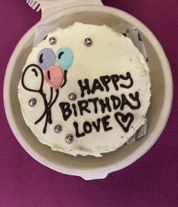 A white-frosted cake in a beige box with pastel balloons and silver pearls, topped with the message “Happy Birthday Love ❤️.