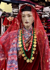 A mannequin dressed in traditional attire featuring a red scarf over a patterned top, adorned with multiple rows of colorful necklaces, including gold coins and bright beads.