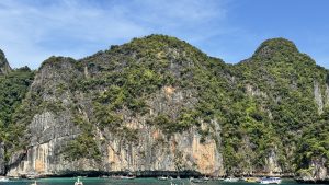 A massive, textured limestone cliff covered in green foliage towering over a line of traditional Thai longtail boats anchored in the emerald sea.