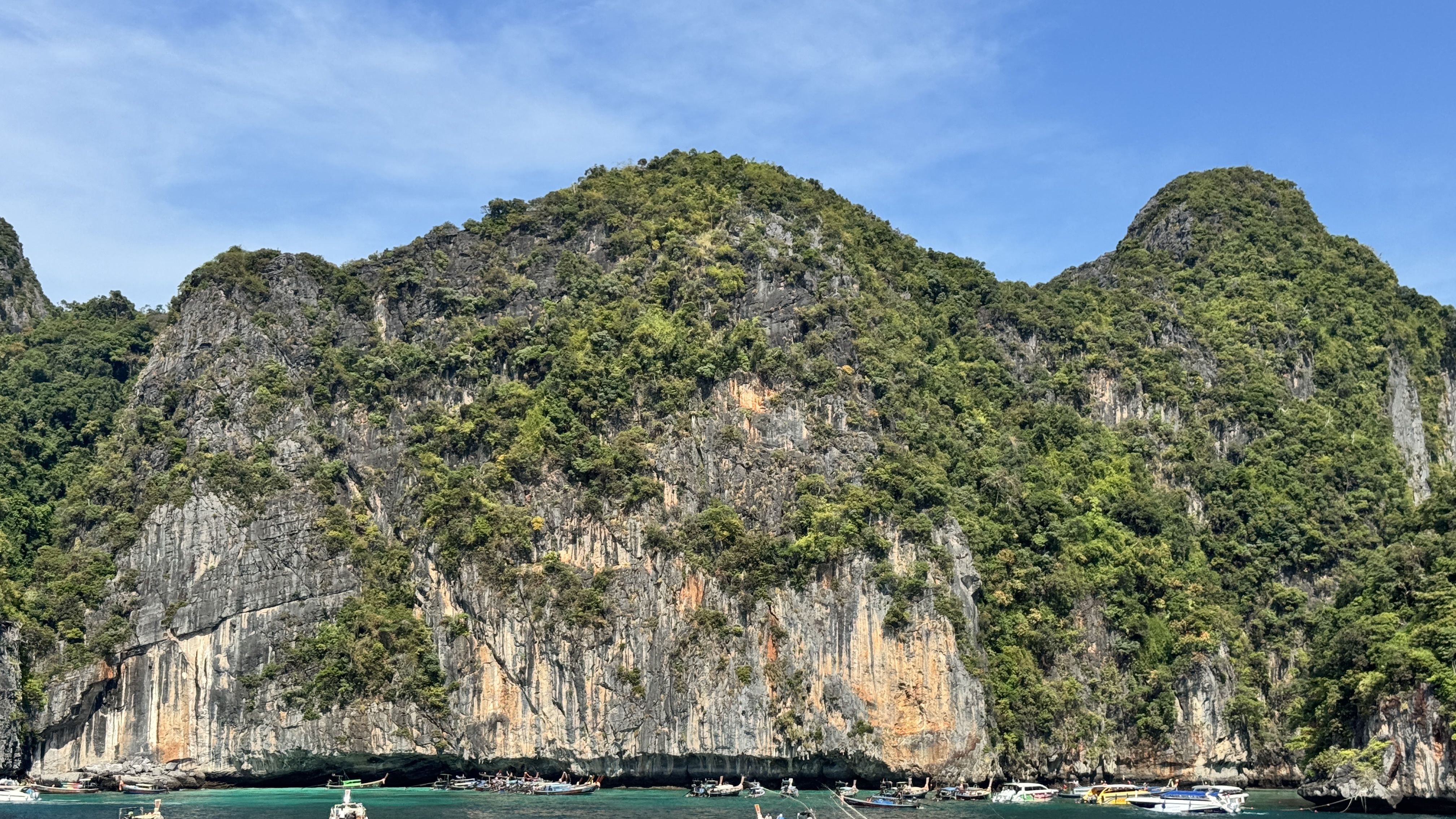 A massive, textured limestone cliff covered in green foliage towering over a line of traditional Thai longtail boats anchored in the emerald sea.