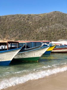 A row of colorful wooden fishing boats, docked on a tropical sandy beach with crystal-clear turquoise water. In the background, a dry forest hill stands under a clear blue sky. The scene captures the essence of a peaceful coastal landscape
