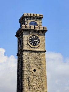 Close-up view of the clock tower at Galle Fort, Sri Lanka, showing the stone structure, clock face with Roman numerals, and crenellated top against a clear blue sky.
