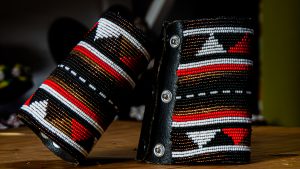 
Two black leather wrist cuffs with detailed white, red, and gold beadwork in geometric patterns rest on a wooden surface.